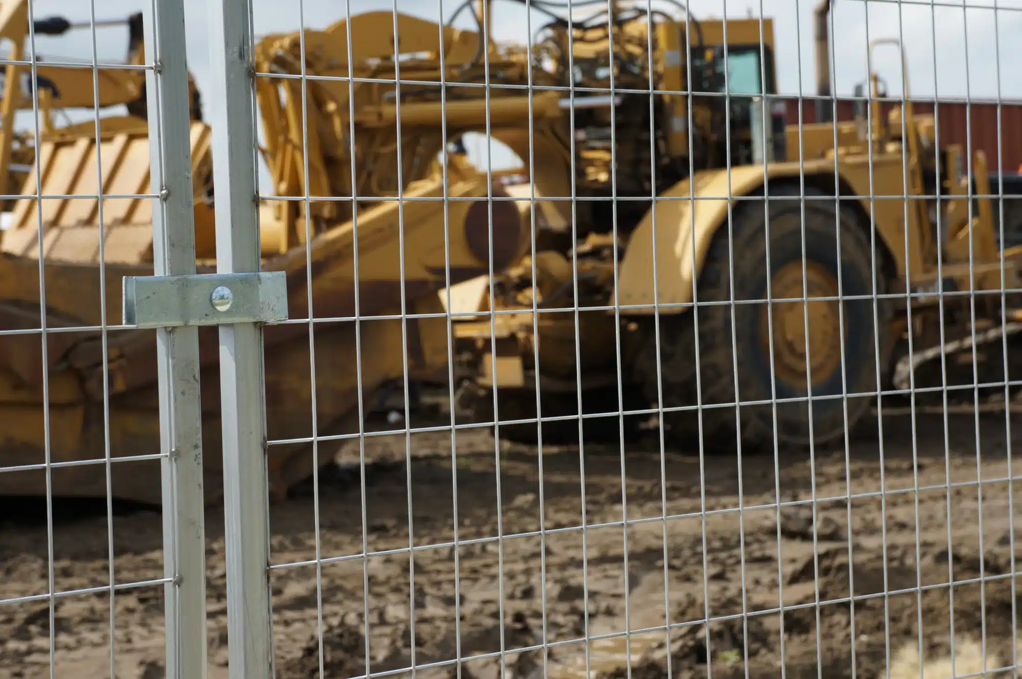 A close up of a construction fence secured by a security clamp, with construction equipment in the background.