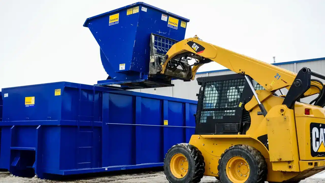 A forklift dumps a tipping hopper into a larger waste bin on a construction site.
