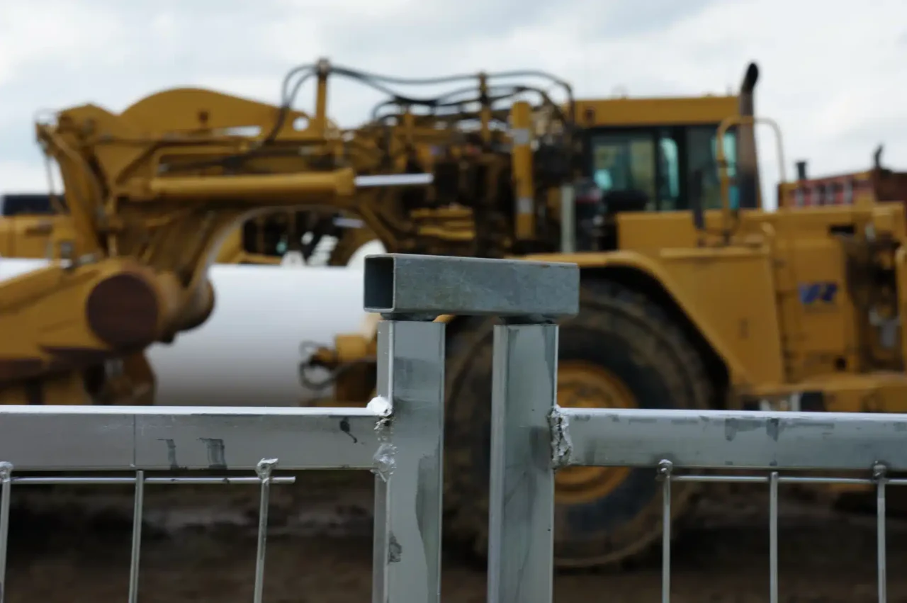 A steel coupler secures two fence panels together in front of heavy equipment.
